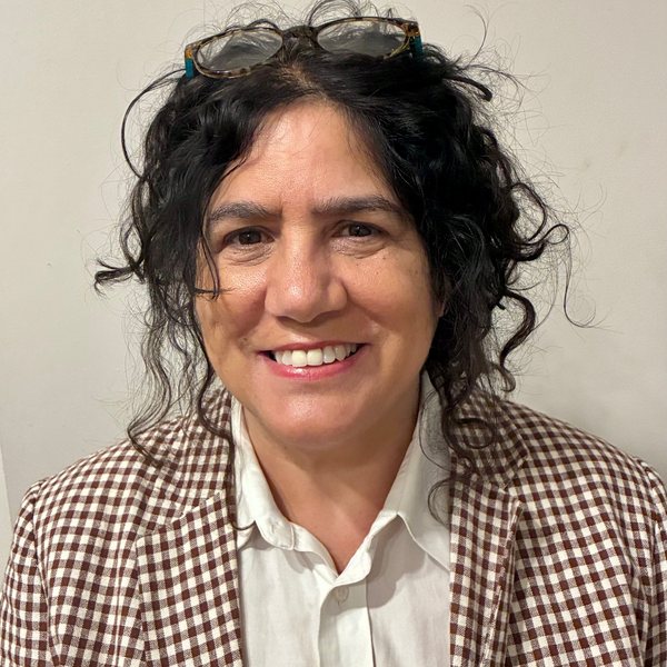 Diane Forte smiling, with curly dark hair and glasses on her head, wearing a brown gingham blazer over a white shirt.