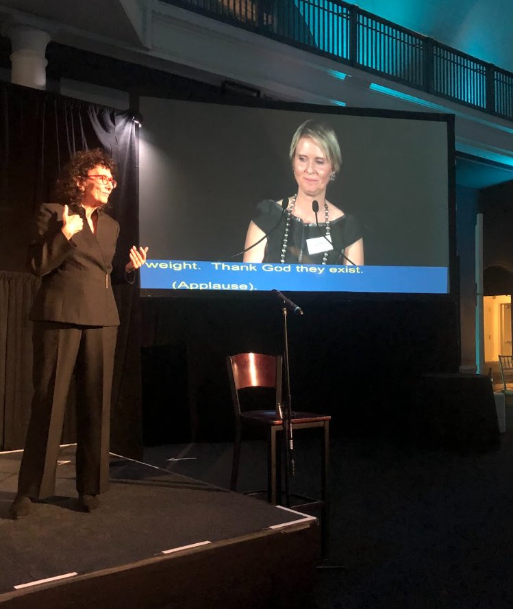 An interpreter signing on stage beside a large screen showing a speaker with live captions.