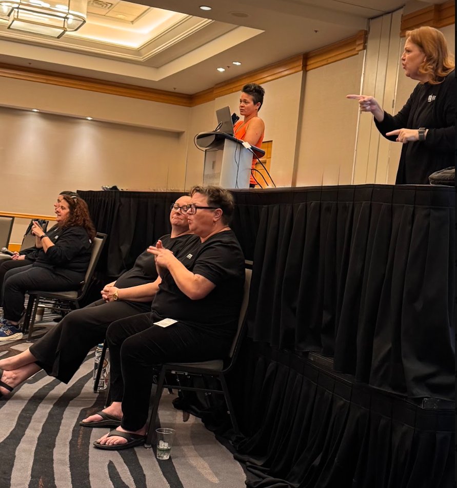 A team of interpreters working together at a conference: one signs from the front of the room while others seated below interpret for participants.