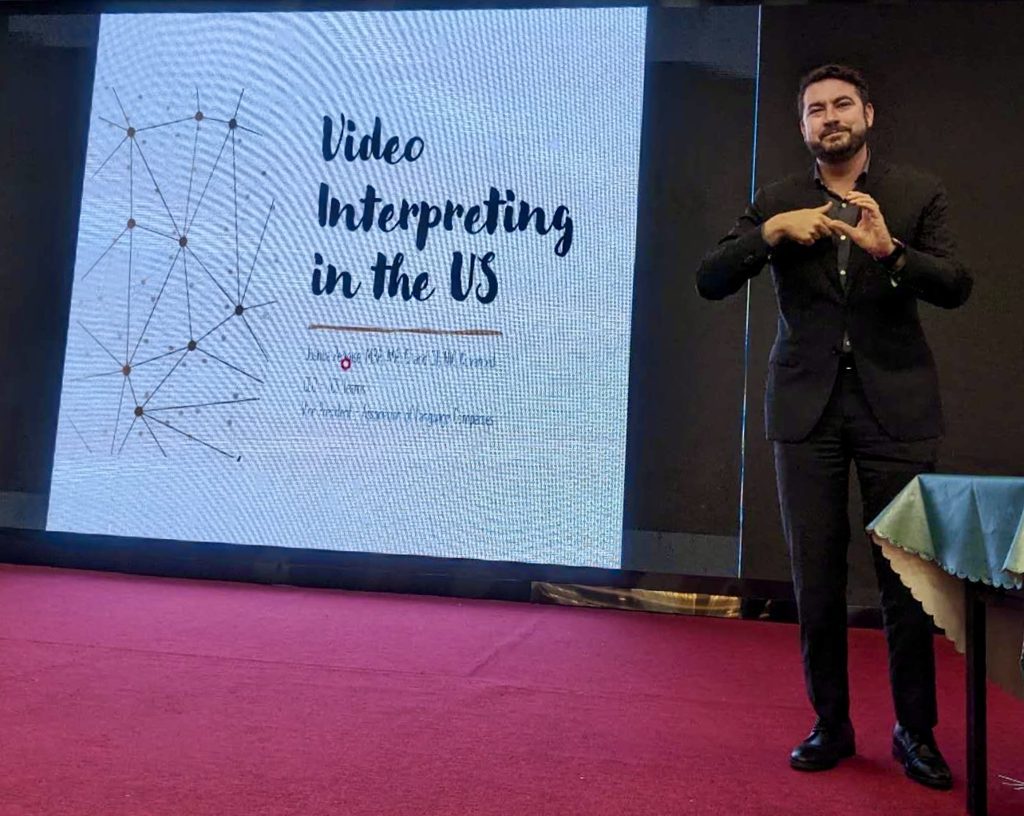 A presenter signing on stage beside a slide titled Video Interpreting in the US.